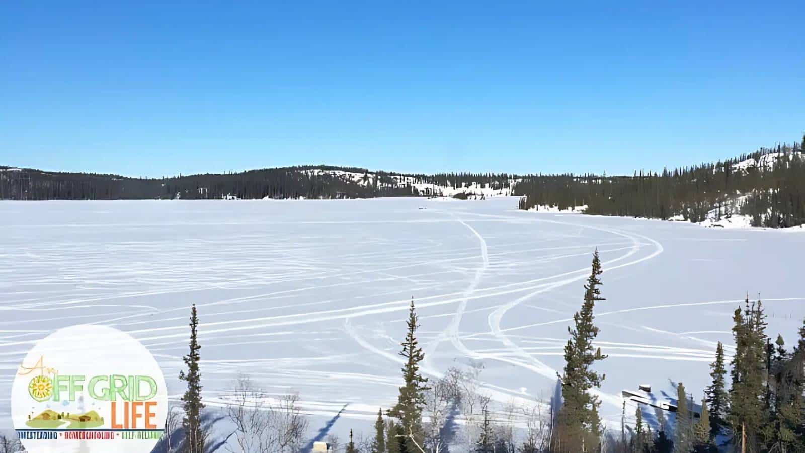 Snow-covered lake with visible snowmobile tracks, surrounded by trees under a clear blue sky. "Off Grid Life" logo at the bottom left corner.