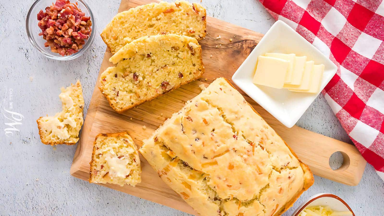 Sliced bacon bread on a wooden board with butter slices on a dish, small bowl of bacon bits, and a red checkered cloth.