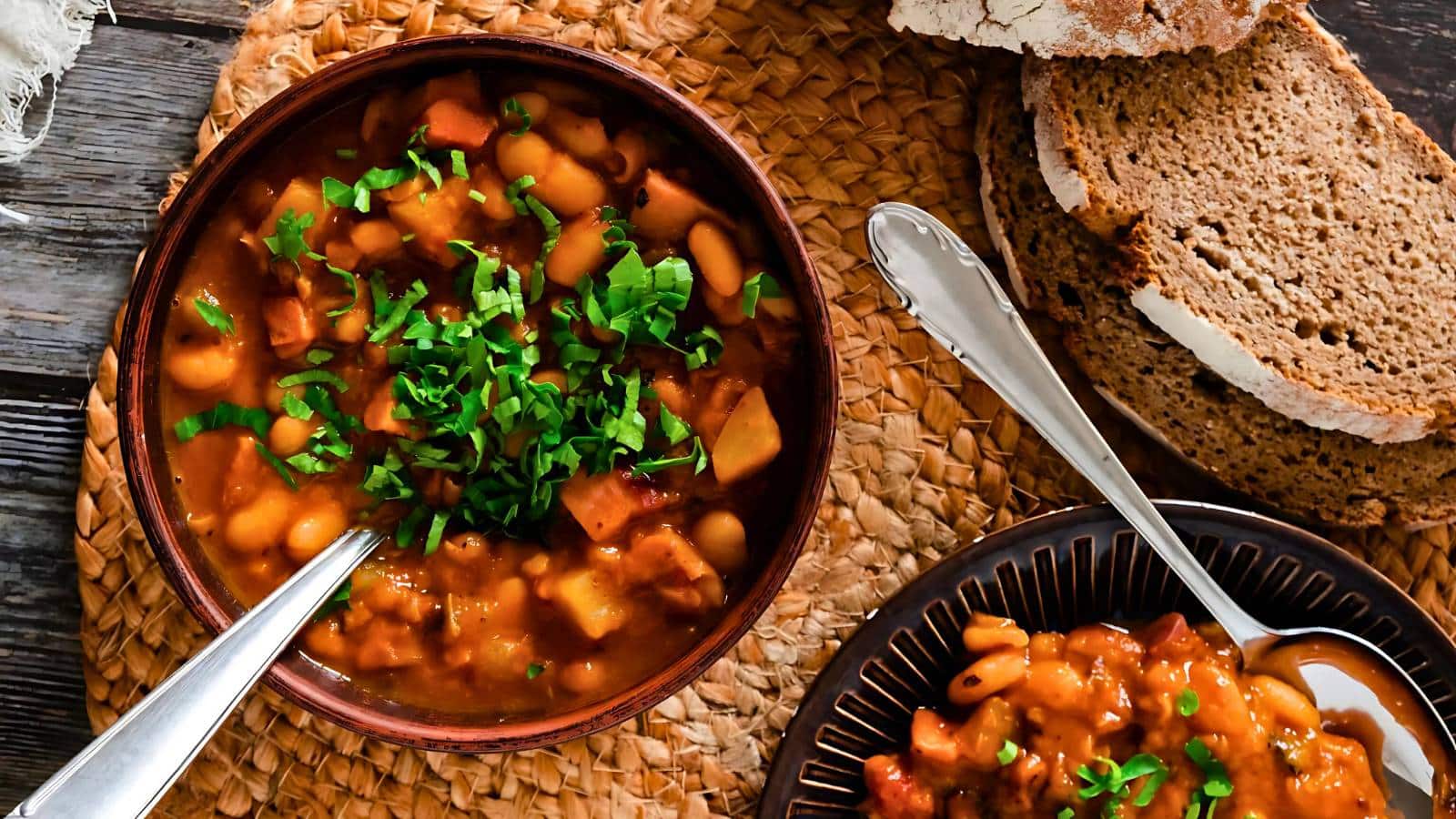 A bowl of bean stew topped with herbs next to slices of brown bread on a woven mat.