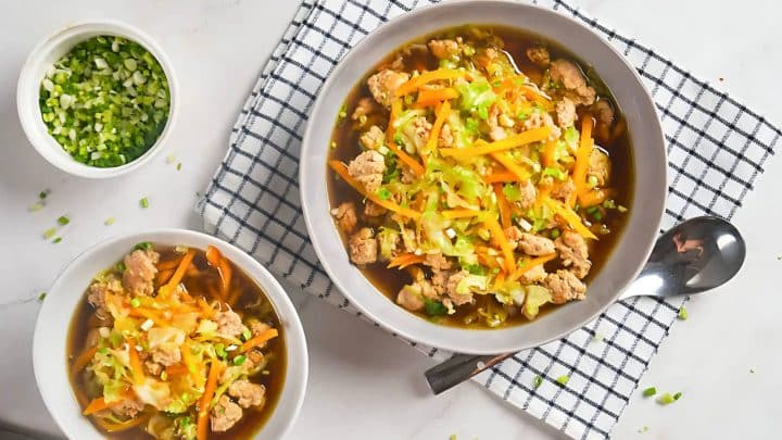 Two bowls of soup with ground meat, shredded carrots, and chopped greens on a checkered cloth. A small bowl of chopped green onions is beside them. A spoon is placed next to the larger bowl.