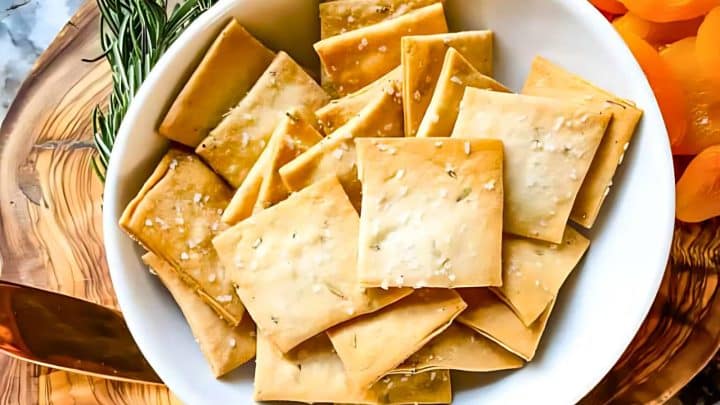 White bowl filled with square crackers, garnished with rosemary, on a wooden surface.