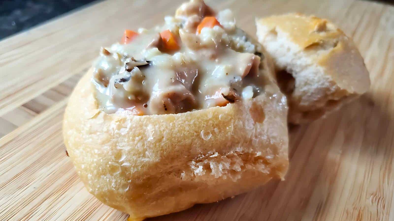 A bread bowl filled with creamy soup containing vegetables and chunks of meat, placed on a wooden surface.
