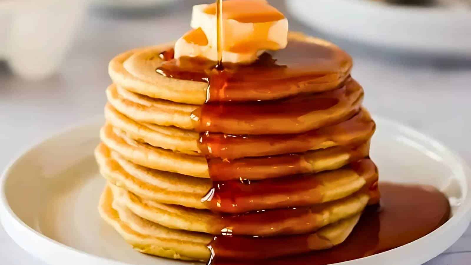 A stack of pancakes topped with butter and syrup on a white plate. Syrup is being poured over the pancakes.