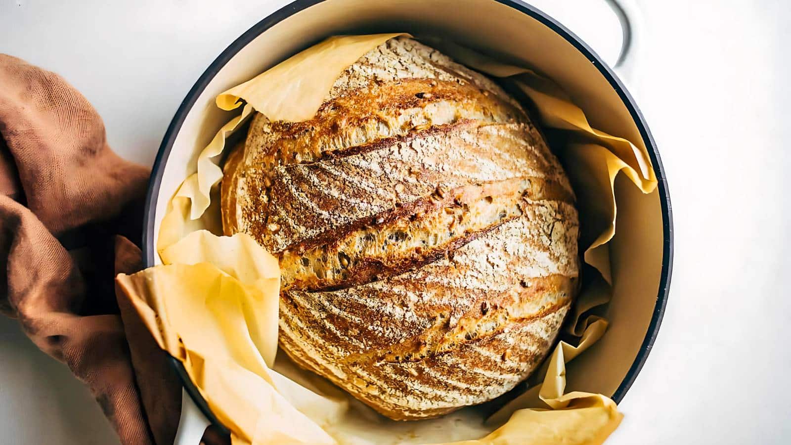 A round loaf of rustic bread with a crusty exterior sits in a parchment-lined dutch oven. A brown cloth is partially visible beside it.