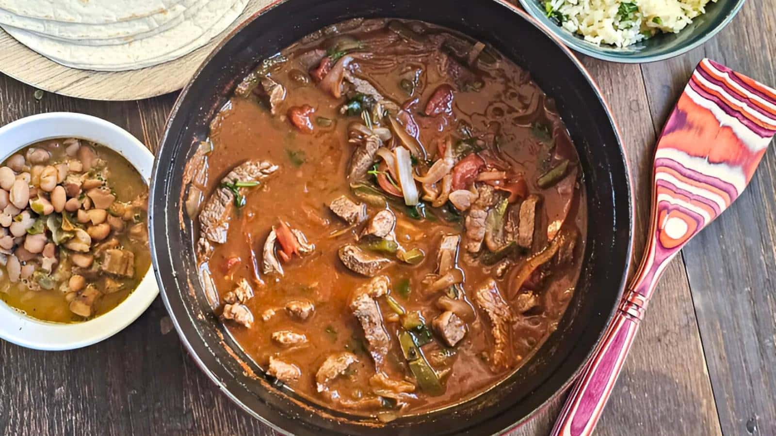 A pot of beef stew with sliced vegetables, a bowl of beans, a plate of tortillas, and a serving spoon on a wooden table.