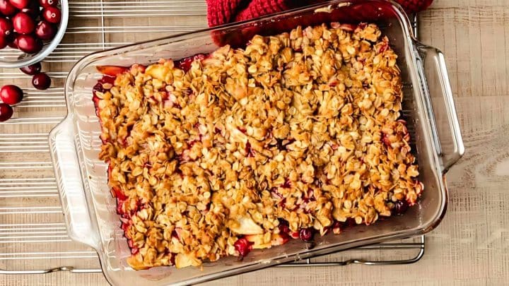 A baked cranberry and apple crisp in a clear glass dish, topped with a golden oat crumble, sits on a metal cooling rack.