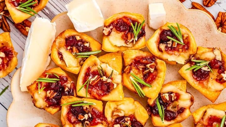 A plate of pastry bites topped with cranberry sauce, nuts, and rosemary, accompanied by slices of soft white cheese.
