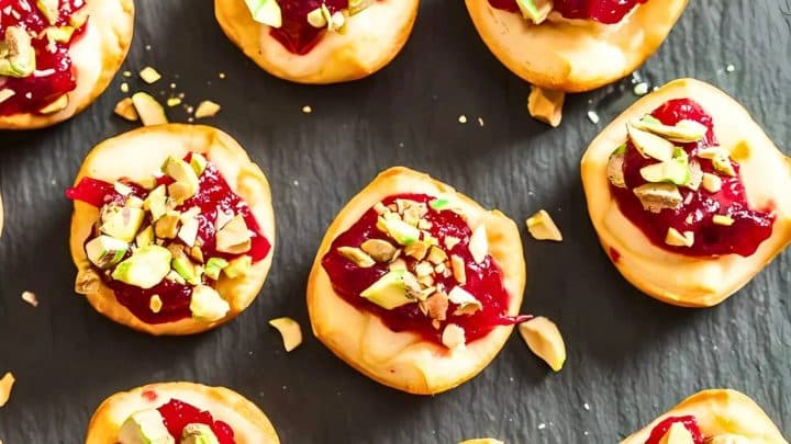 Mini pastries topped with red jam and chopped pistachios on a dark slate surface.