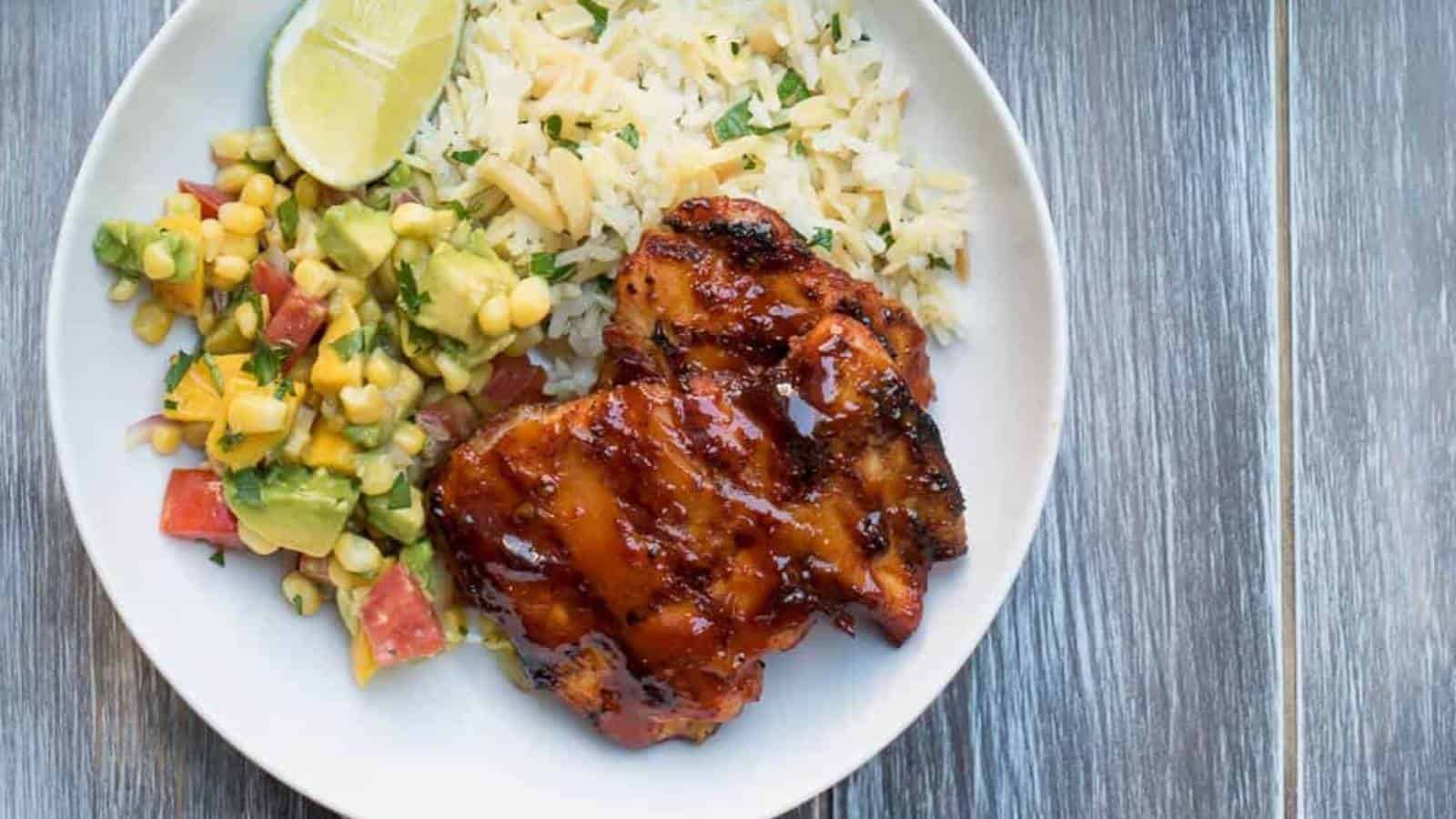 A plate with grilled chicken, white rice garnished with herbs, a lime wedge, and a side of corn, avocado, and tomato salad on a wooden surface.
