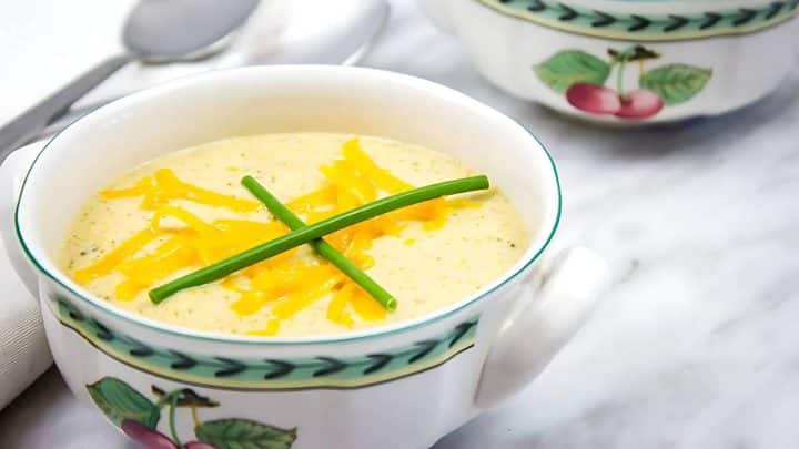 Soup in a decorative bowl with shredded cheese and chives on top, on a marble surface with another bowl partially visible.