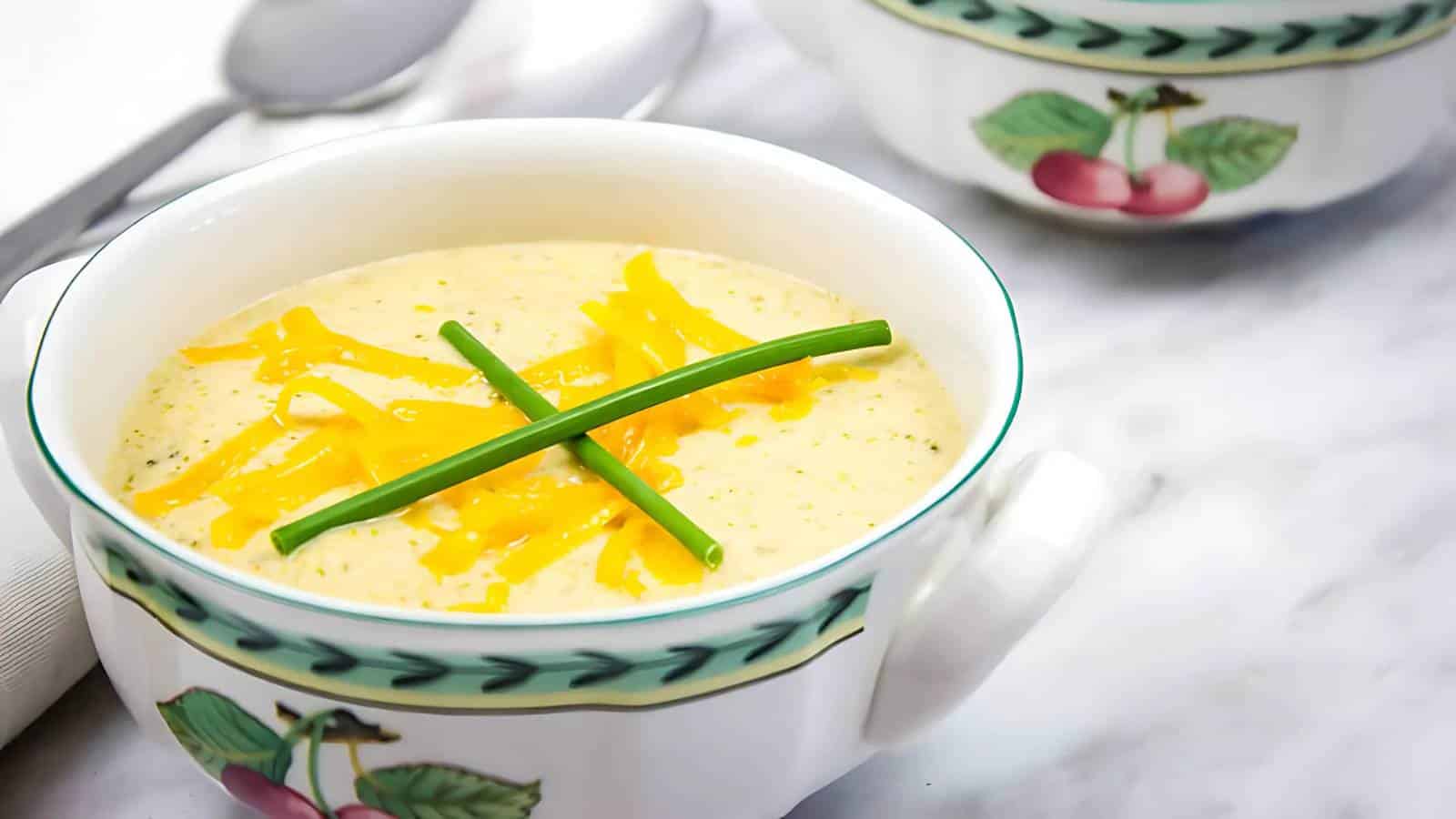 Soup in a decorative bowl with shredded cheese and chives on top, on a marble surface with another bowl partially visible.