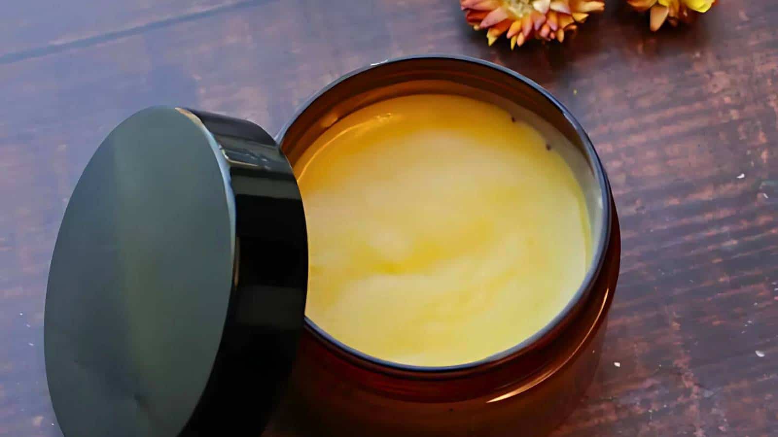 An open jar of yellow ointment sits on a wooden surface, with dried flowers nearby.