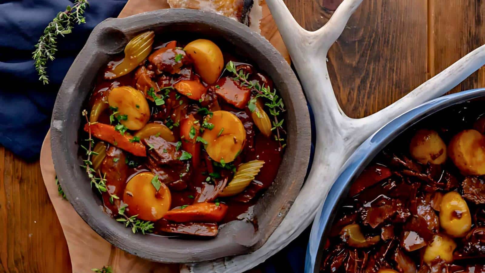 A bowl of beef stew with carrots, potatoes, and herbs on a wooden surface, next to a blue pot containing more stew.