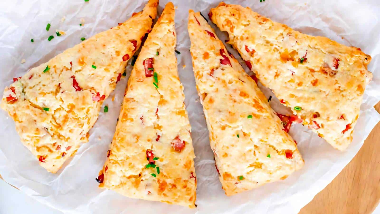 Four triangular cheese and bacon scones on parchment paper, garnished with chopped chives.