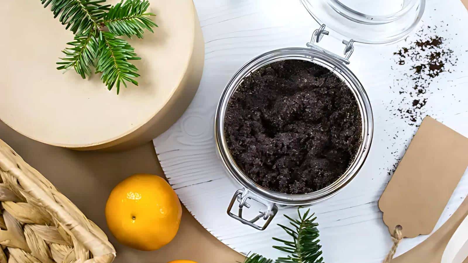 A jar of dark scrub sits open on a white surface, surrounded by pine branches, oranges, a circular box, and a woven basket. A blank brown tag is nearby.