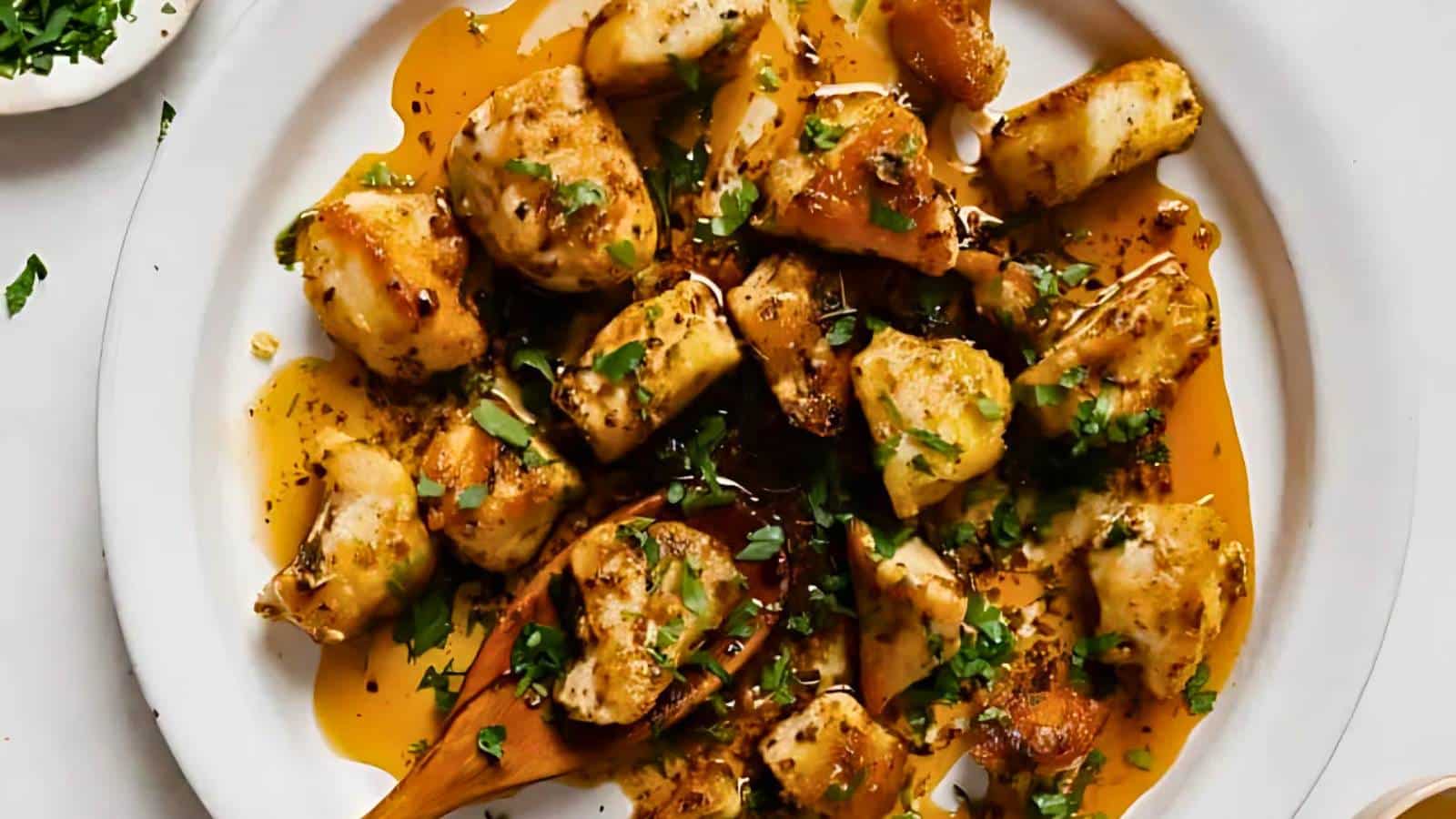A plate of chicken pieces glazed with a thick, orange sauce, garnished with green herbs, with a wooden spoon resting on the plate.