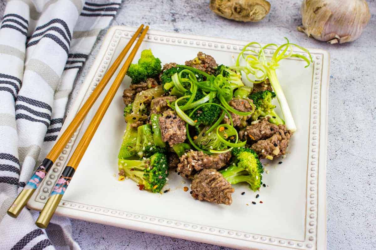 A square white plate with beef and broccoli stir-fry garnished with green onions. Chopsticks and a striped napkin are beside the plate. Garlic and ginger are visible in the background.
