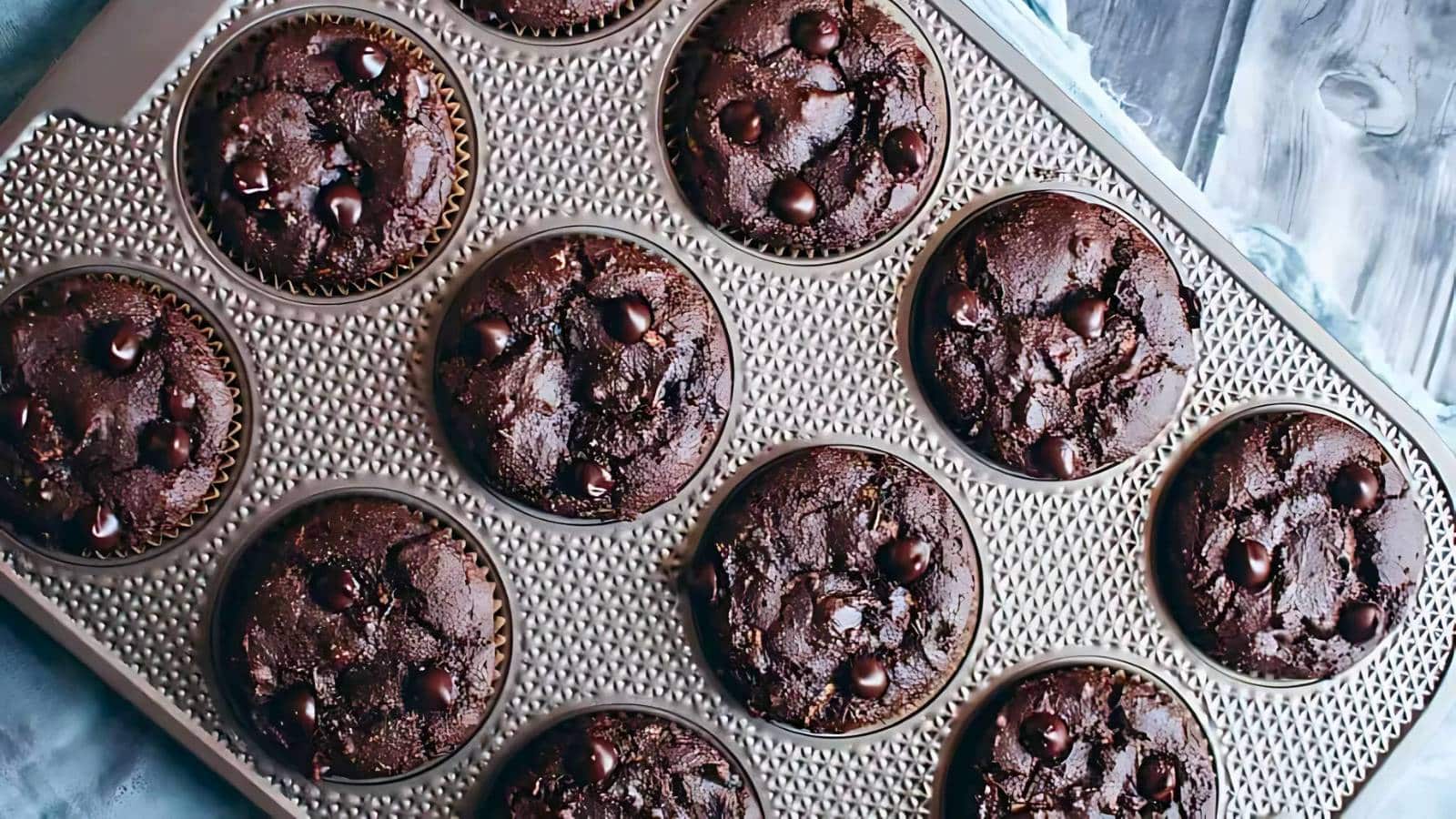 A stack of three chocolate muffins in paper wrappers is set against a blue background, with scattered chocolate chips nearby.