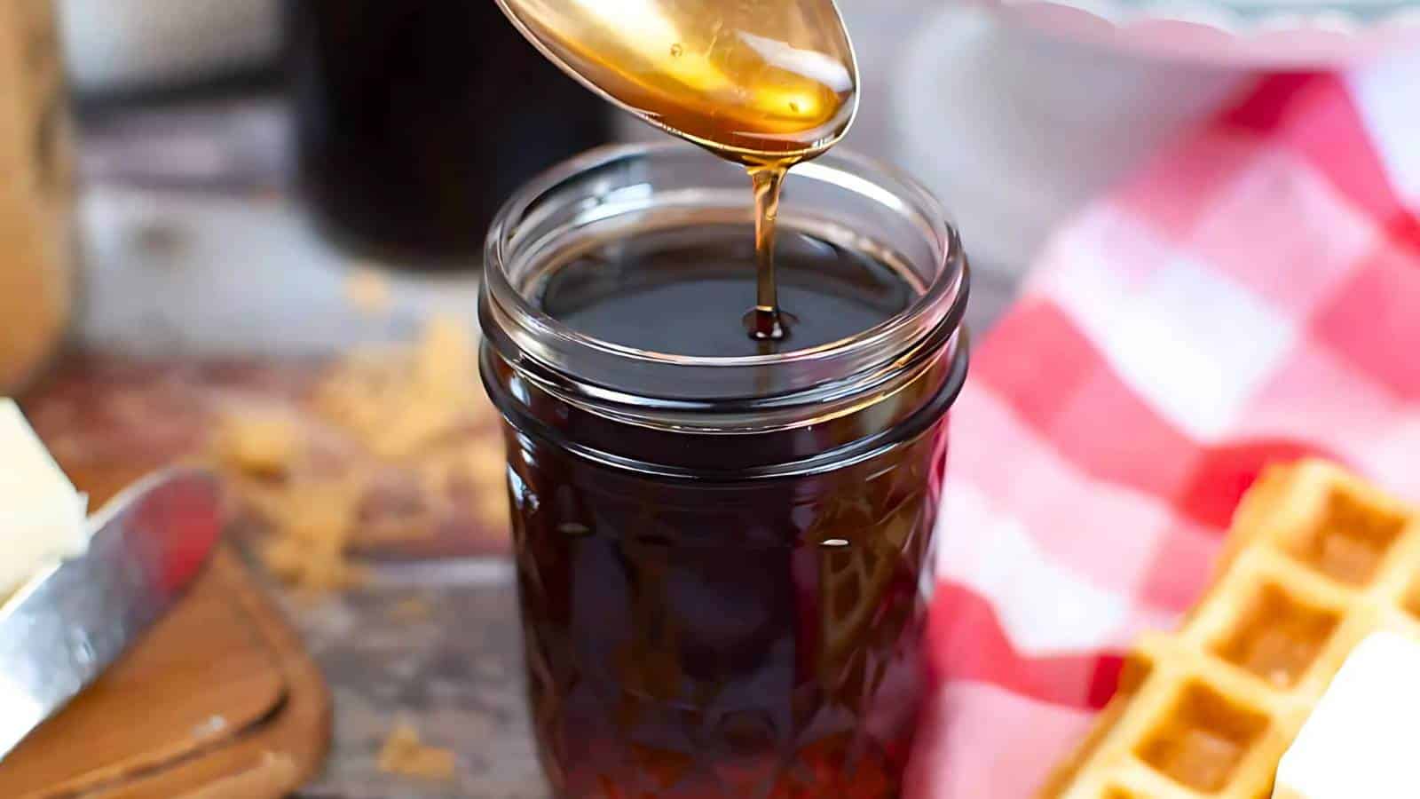 A spoon drizzles syrup into a glass jar next to waffles and a red checkered cloth.