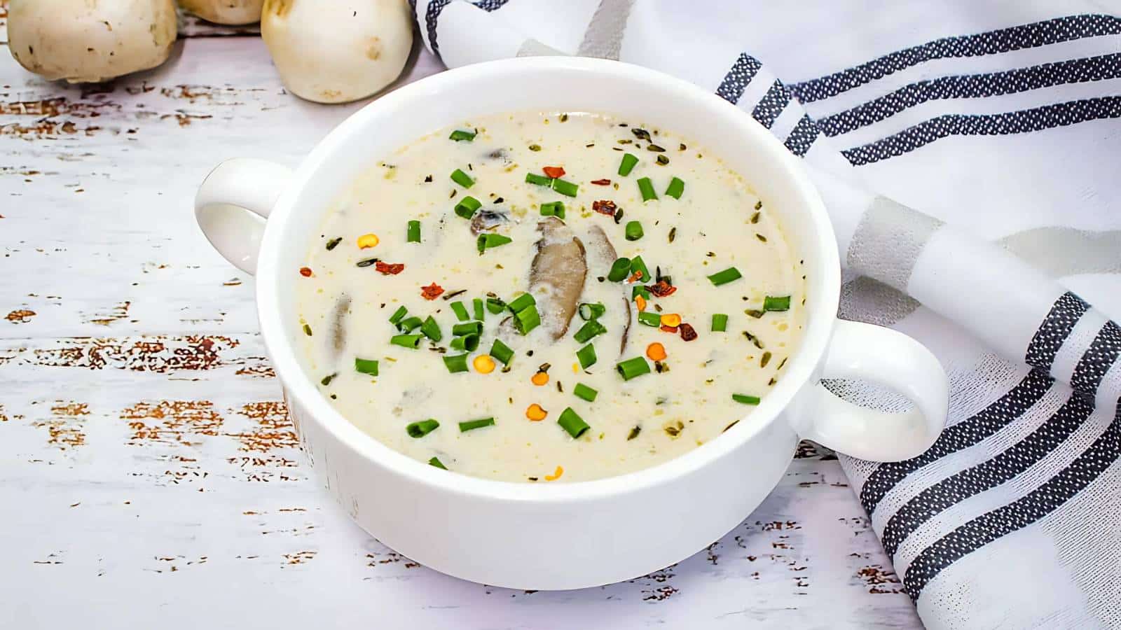 A white bowl of creamy mushroom soup garnished with chopped chives and red pepper flakes, placed on a rustic wooden table beside a striped cloth.