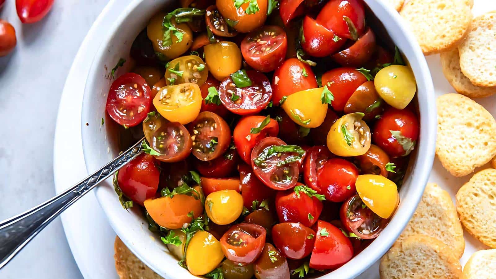 A bowl of mixed colorful cherry tomatoes, chopped and garnished with herbs, with a spoon inside. Slices of bread are placed around the bowl.