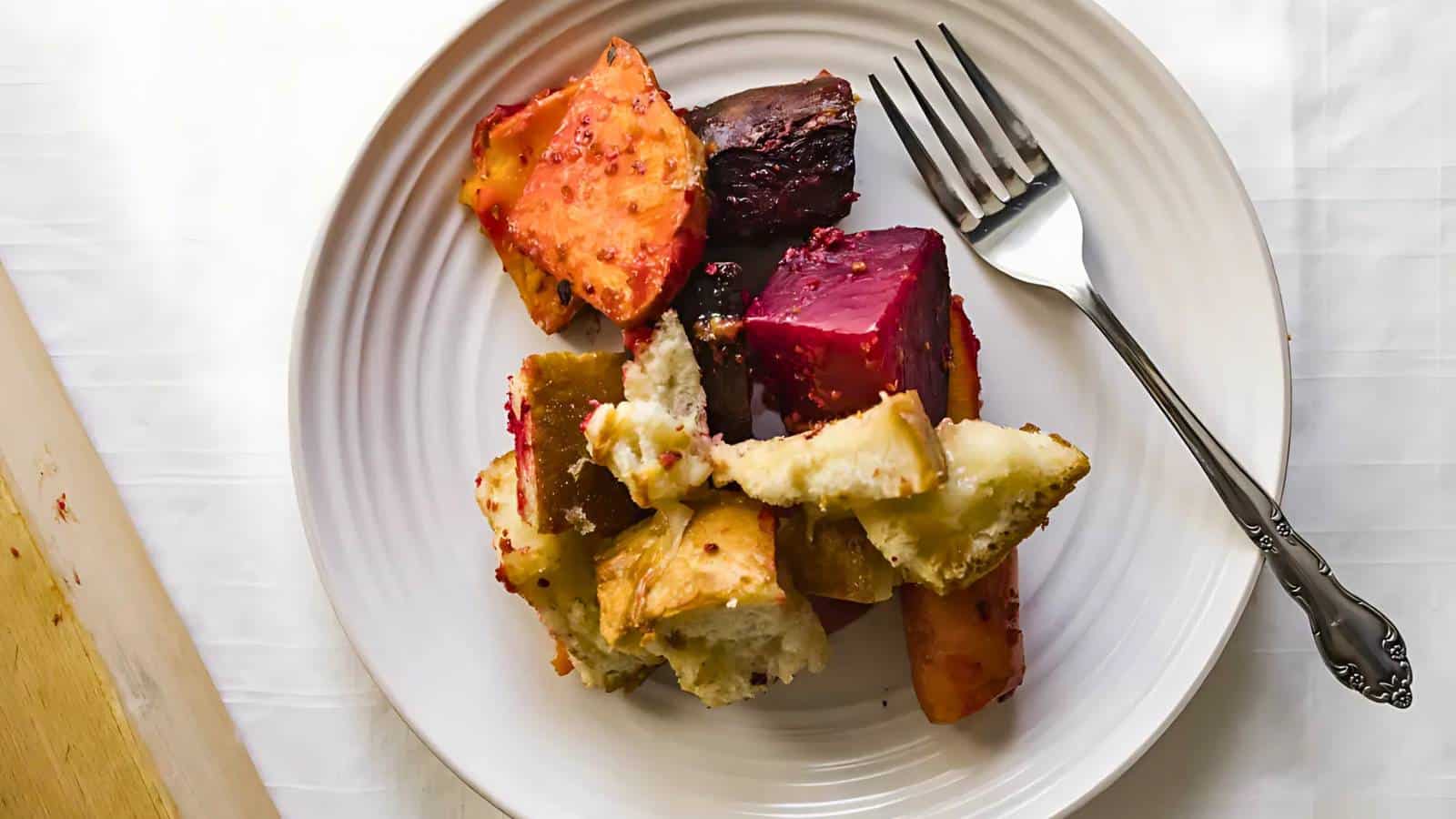 A white plate with assorted roasted vegetables and bread chunks, accompanied by a silver fork on the right.