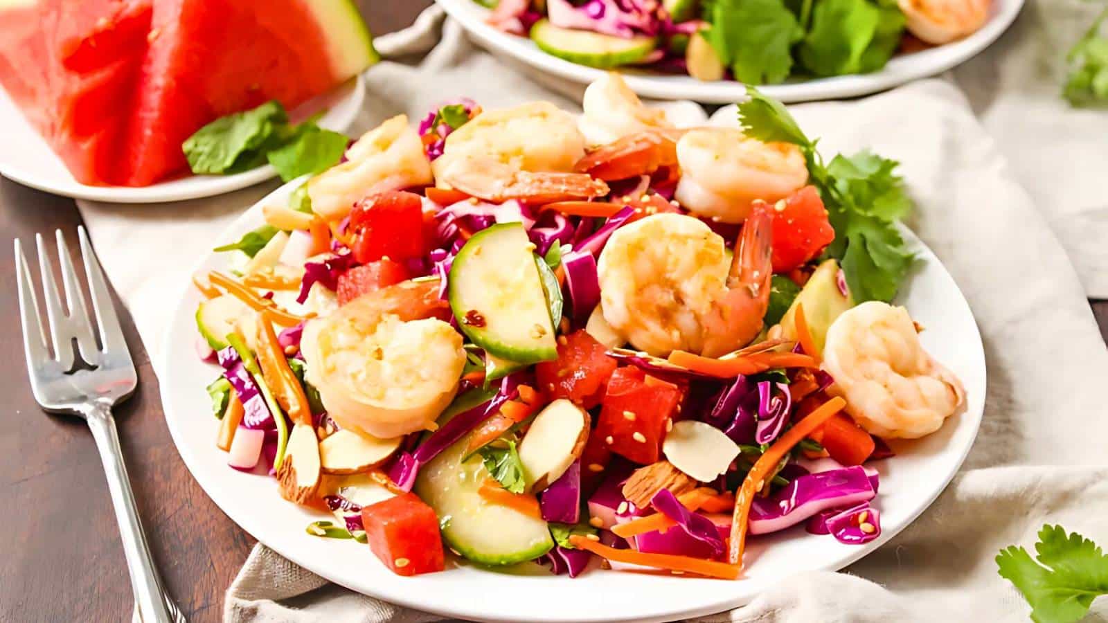 A plate of shrimp salad with cabbage, cucumber, carrots, tomatoes, and almonds, garnished with cilantro, on a wooden table with a fork and watermelon slice nearby.