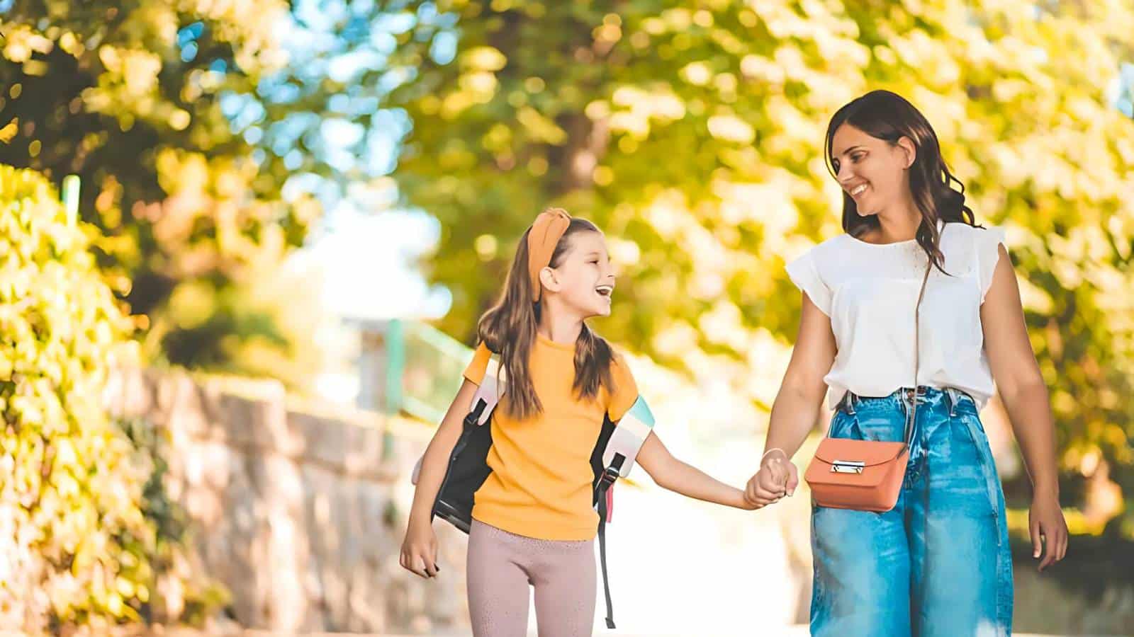 A woman and a girl holding hands while walking outdoors on a sunny day, with trees in the background. The girl has a backpack, and both are smiling.