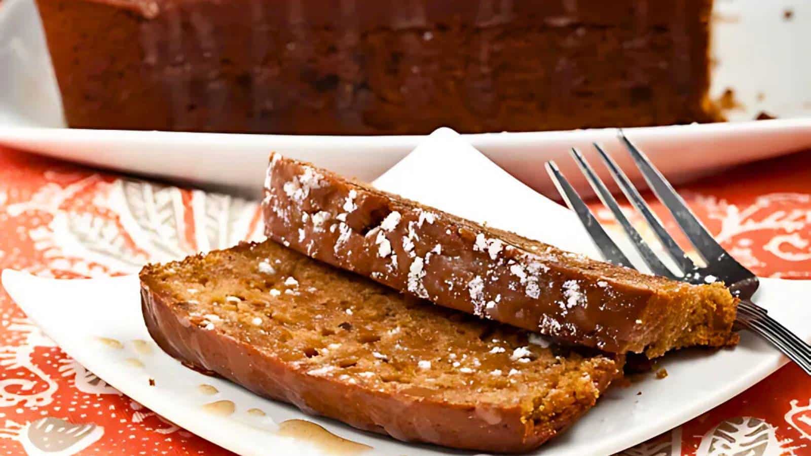 Two slices of spiced loaf cake, dusted with powdered sugar, are served on a white plate with a fork on the side. The tablecloth beneath is red with a white floral pattern.