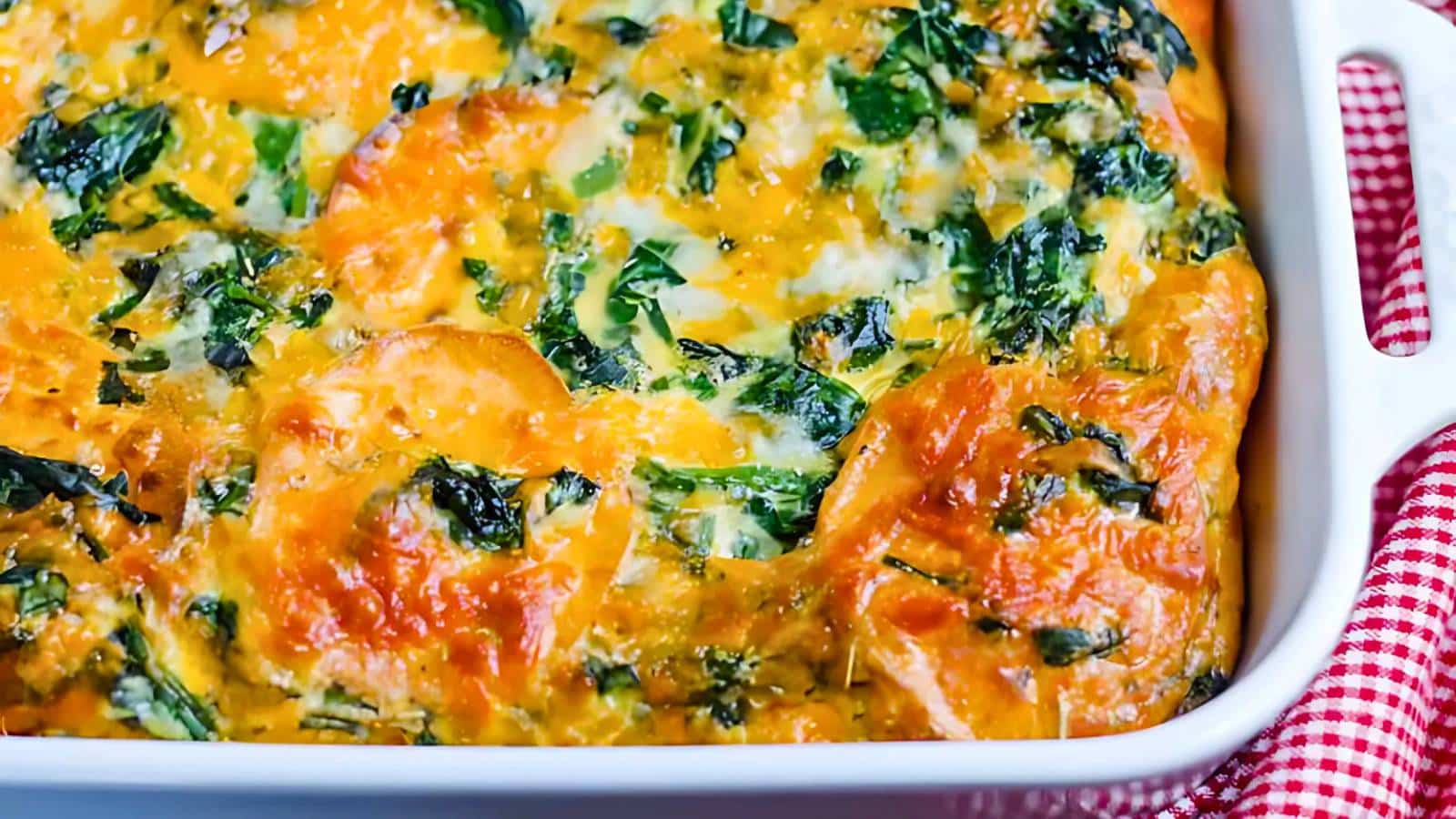 Close-up of a baked vegetable casserole in a white dish, topped with golden-brown cheese and herbs. A red and white checkered cloth is partially visible in the background.