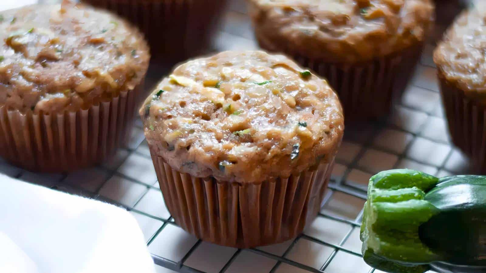 Several zucchini muffins on a cooling rack with a sliced zucchini nearby.