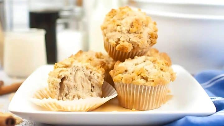 A plate of four crumb-topped muffins, with one muffin partially unwrapped, is displayed on a white square plate.