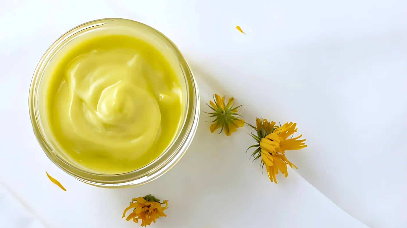A jar of yellow cream next to three small yellow flowers on a white surface.