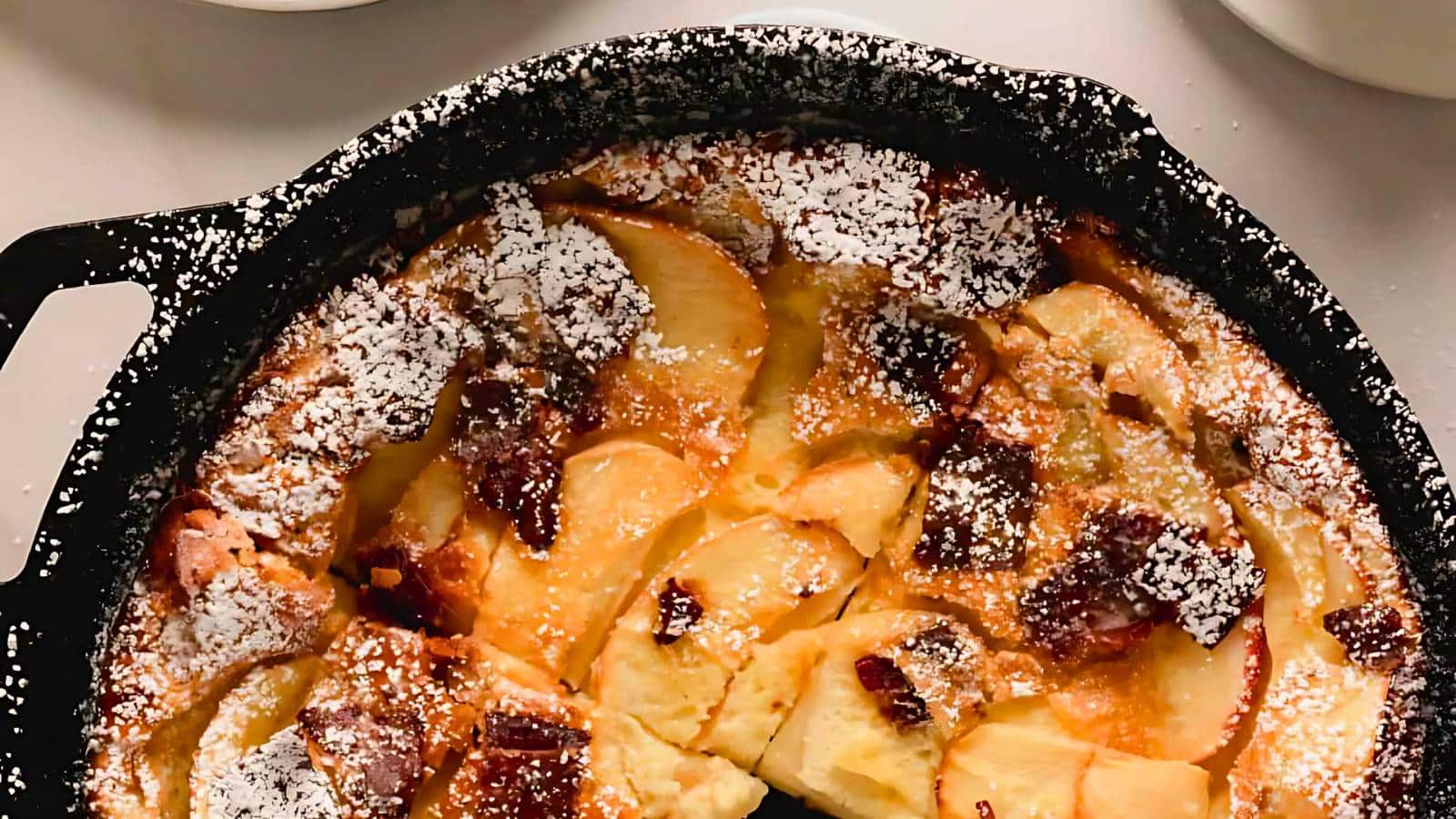 A baked apple and bread pudding in a round black skillet, dusted with powdered sugar.
