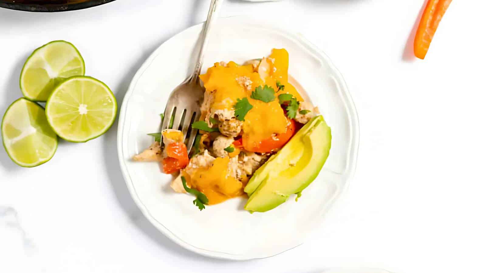A plate of cheesy chicken mixture with tomatoes and herbs, garnished with avocado slices. Lime wedges are on the side.