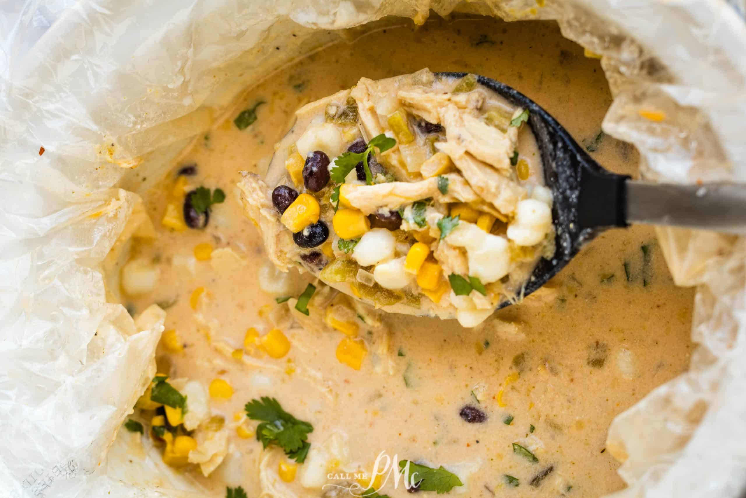 A creamy soup with corn, black beans, chicken, and herbs is being ladled from a pot lined with a cooking bag.