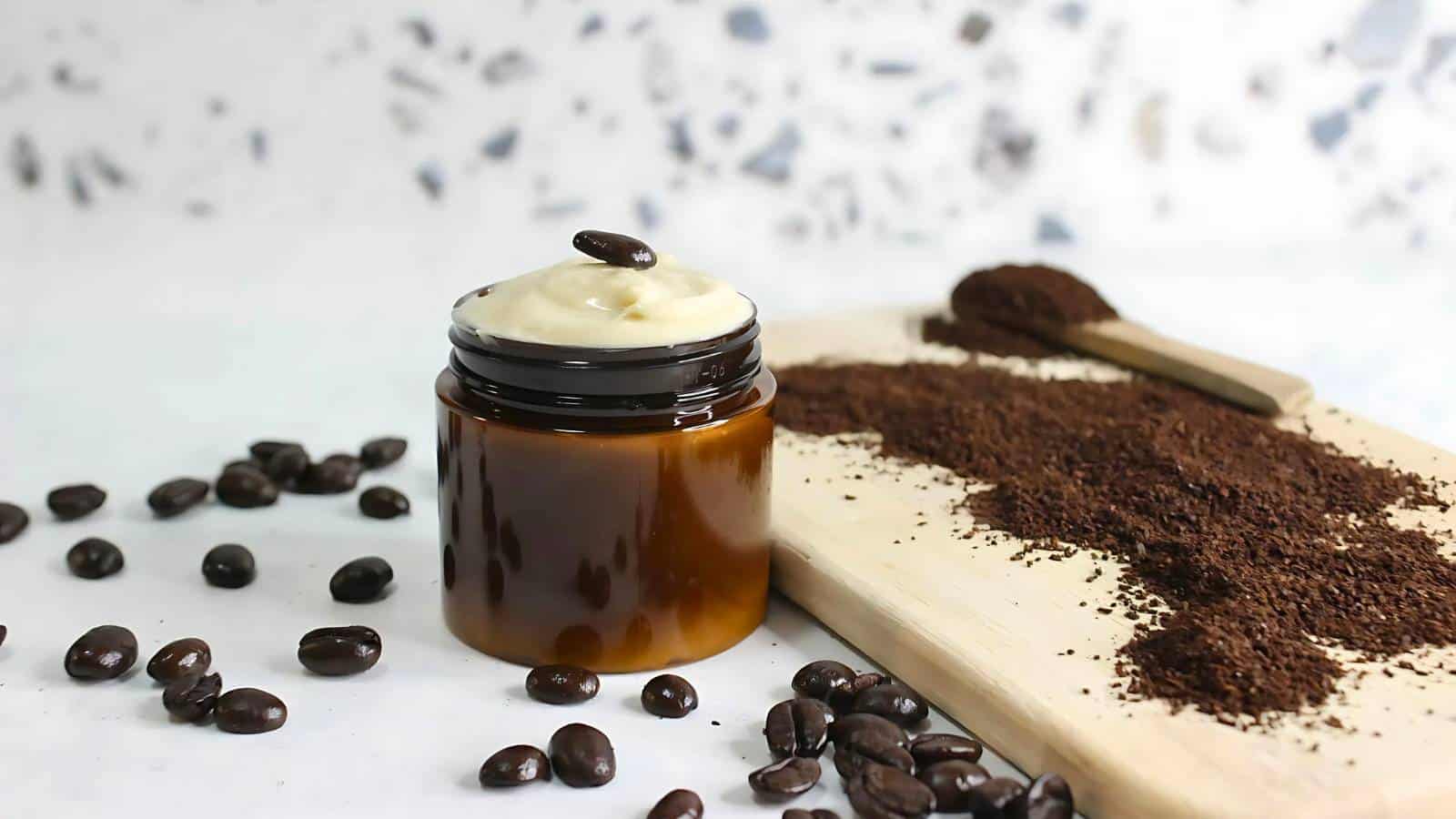 A jar of creamy substance topped with a coffee bean, surrounded by scattered coffee beans and a wooden board with ground coffee.