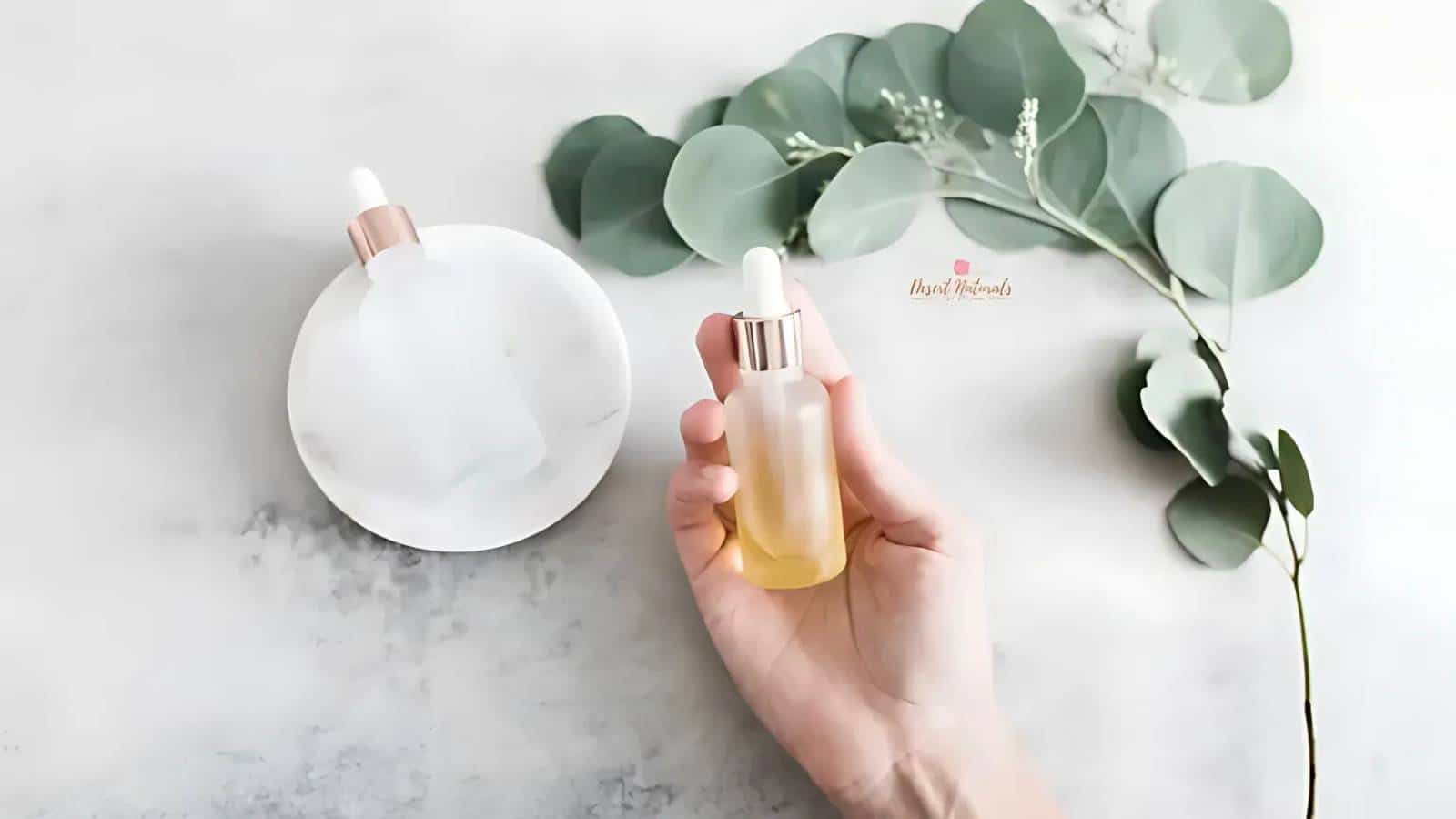 A hand holds a dropper bottle of yellow liquid. Another bottle rests on a white dish. Eucalyptus leaves are arranged nearby on a light surface.