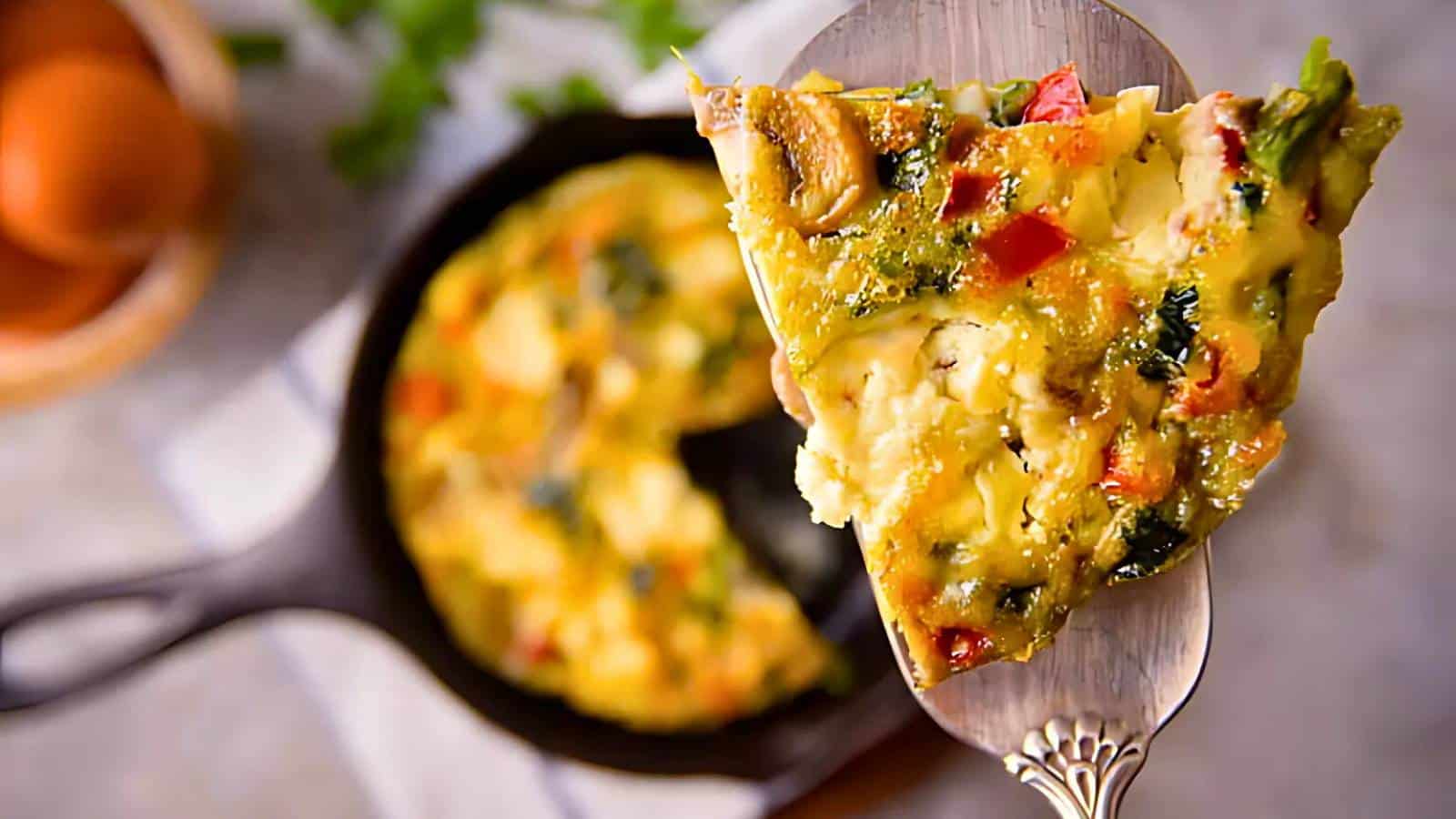 Slice of vegetable frittata with mushrooms, spinach, and red peppers held above a skillet.