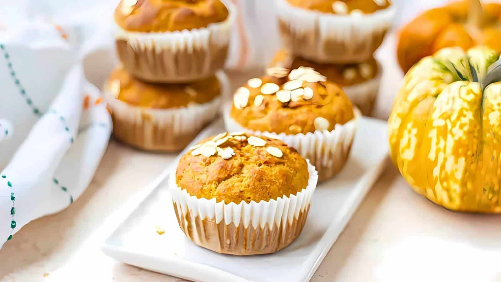 A plate of muffins with oats on top, surrounded by a small pumpkin and a stack of more muffins in the background.