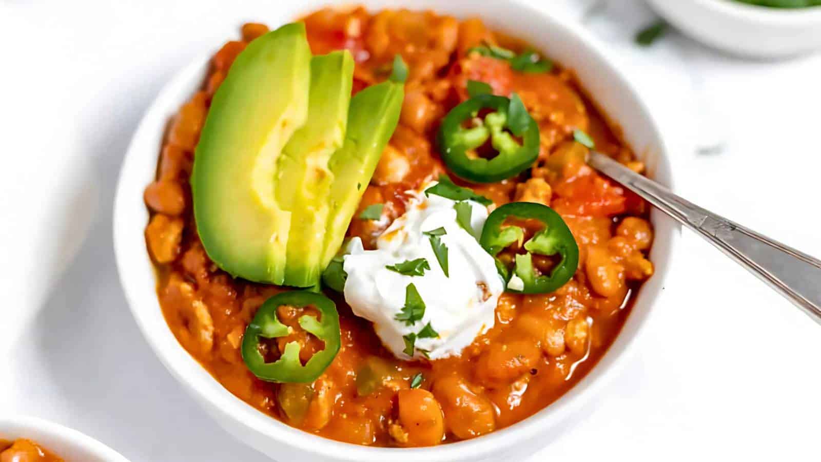 A bowl of chili topped with sliced avocado, jalapeños, sour cream, and cilantro. A spoon rests in the bowl.