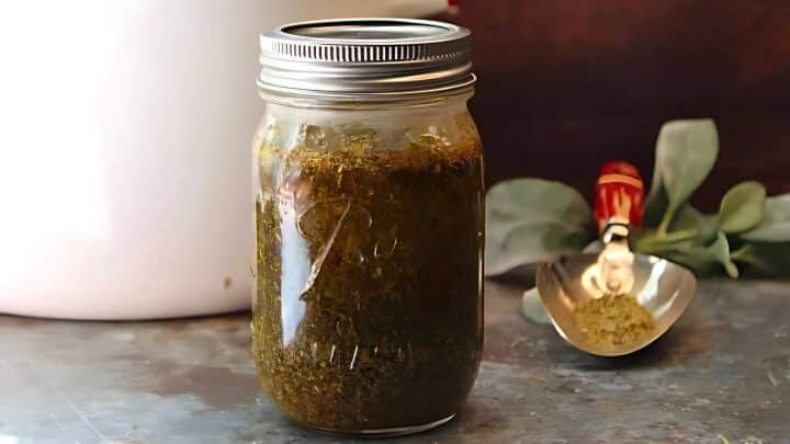 A glass jar filled with green pesto sauce on a kitchen counter, with a metal scoop and fresh herbs in the background.