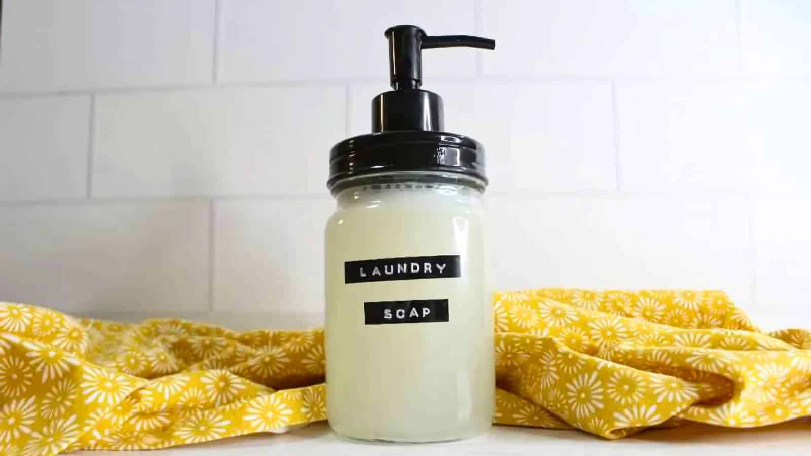 Plastic spoon with blue liquid over a small container. Text on fabric reads "Dreams Est'd 1850 Paradise, Let's Go to California." Yellow fabric partially visible in the background.