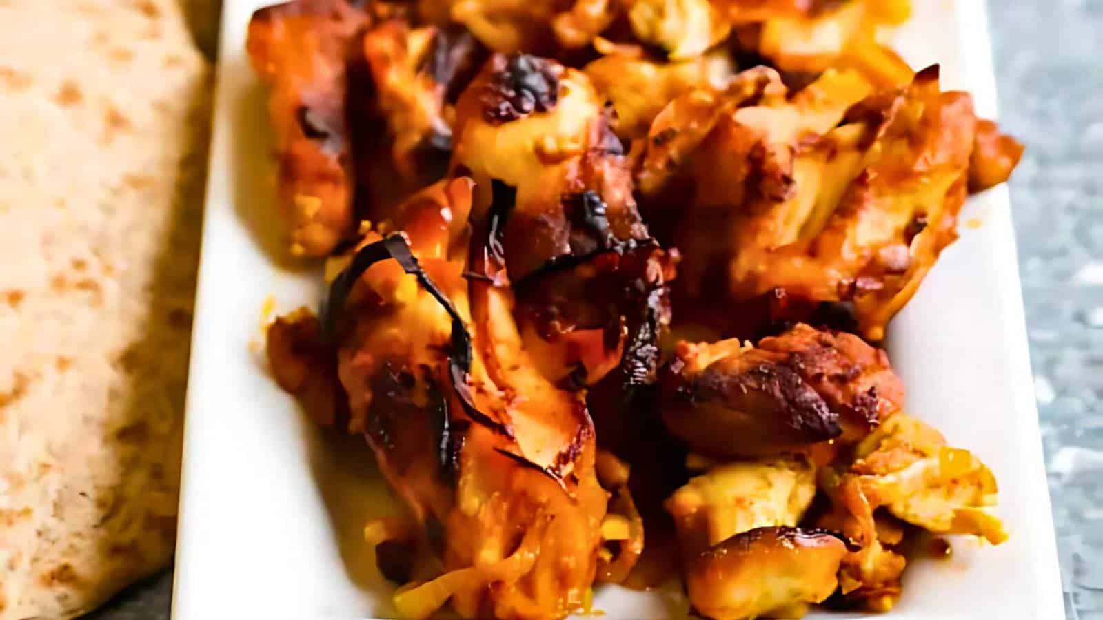 A plate of grilled, marinated chicken pieces with a crispy, charred exterior, next to flatbread.