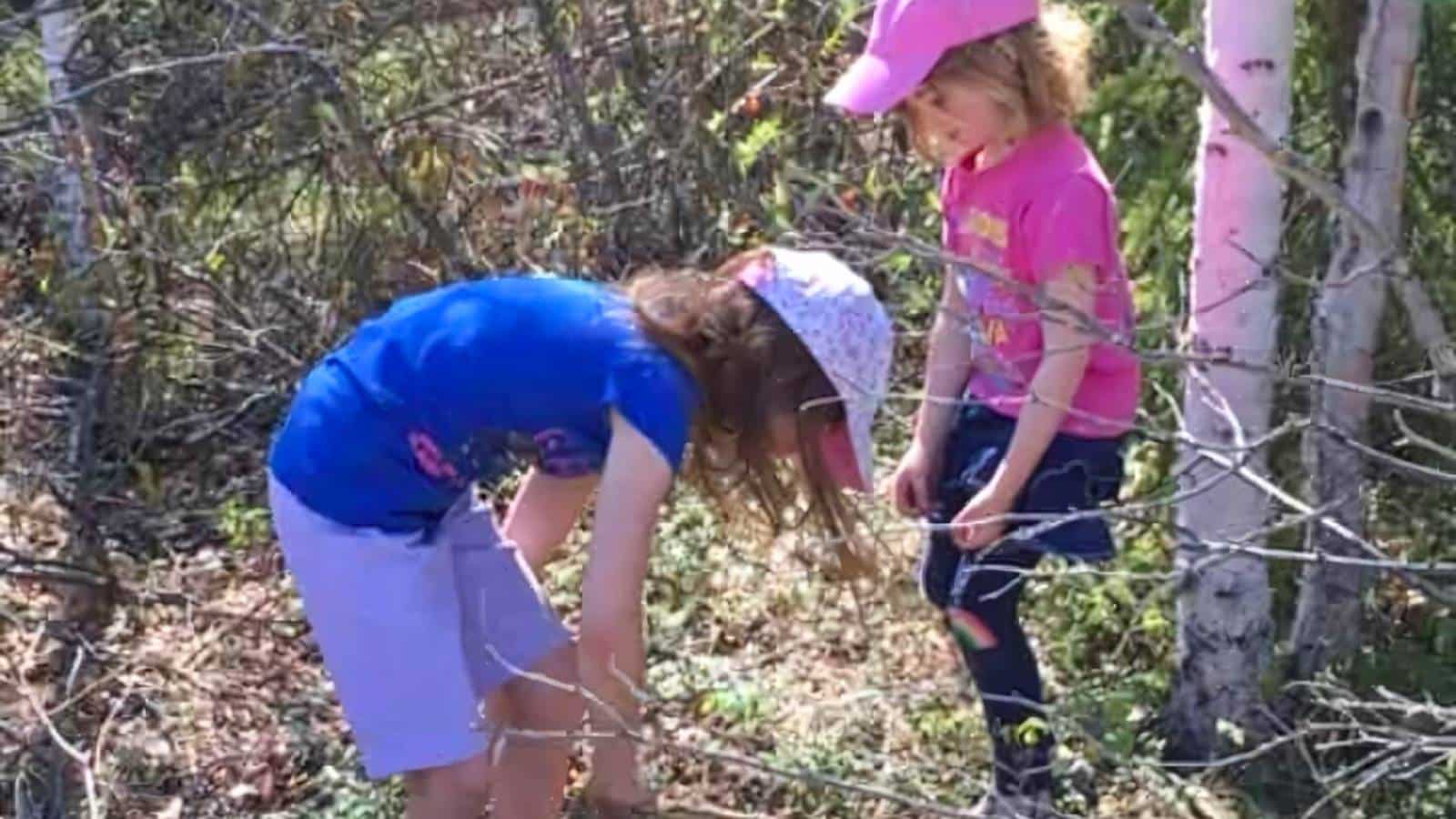 Two children wearing hats are exploring in a forested area. One child is bending down looking at the ground while the other stands nearby, holding a stick.