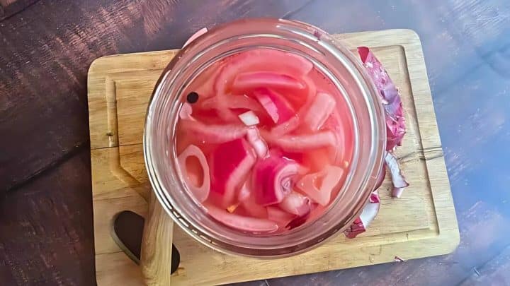 A glass jar filled with sliced red onions in pink liquid on a wooden board with a knife and onion peels around it.