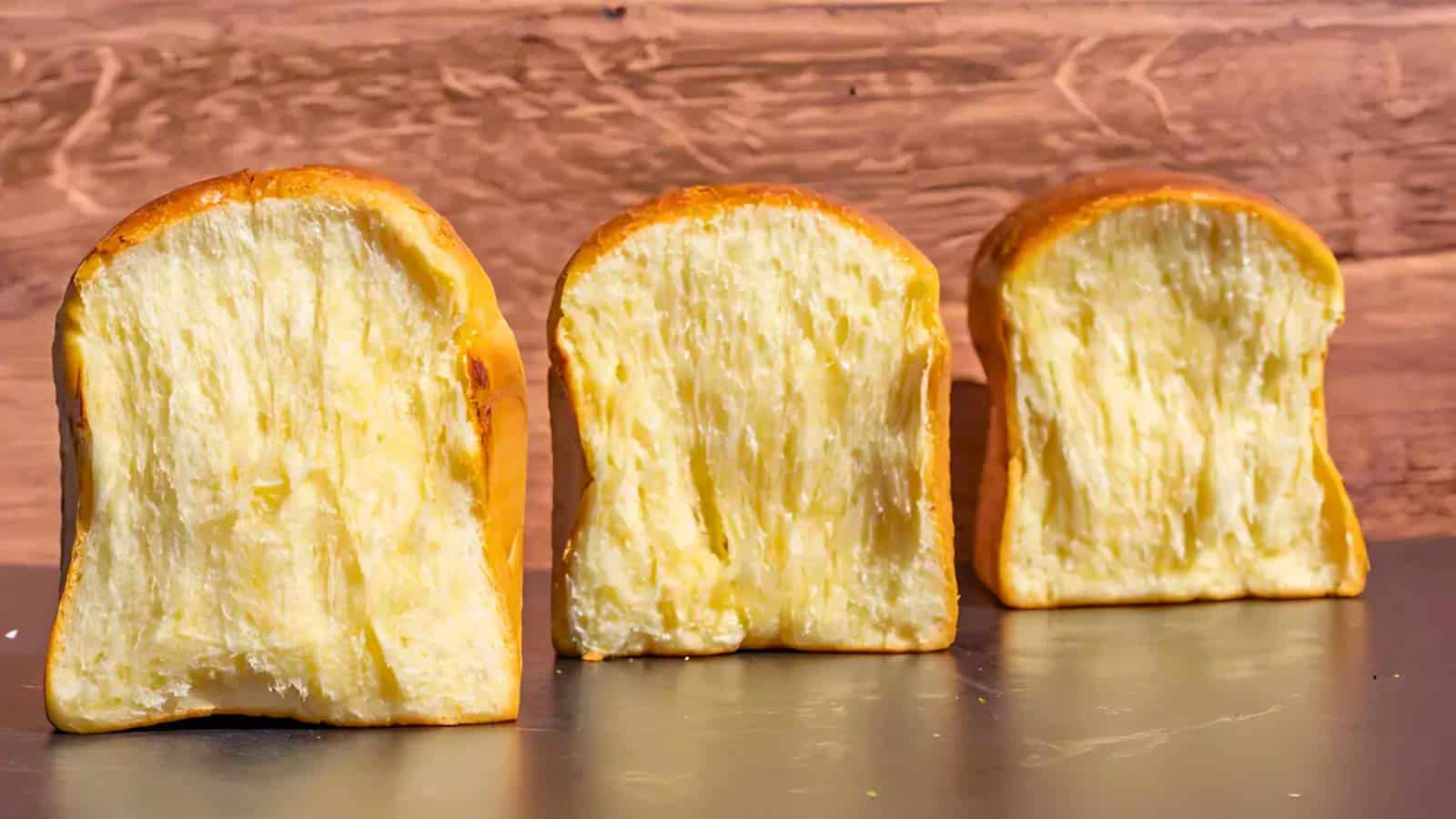 Three slices of fluffy, golden bread are displayed on a dark surface against a wooden background.