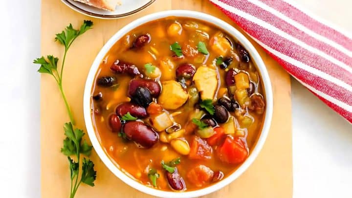 A bowl of hearty vegetable and bean soup on a wooden board, garnished with parsley. A slice of bread and a red-striped napkin are beside it.