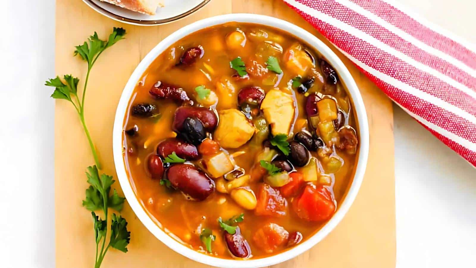 A bowl of hearty vegetable and bean soup on a wooden board, garnished with parsley. A slice of bread and a red-striped napkin are beside it.