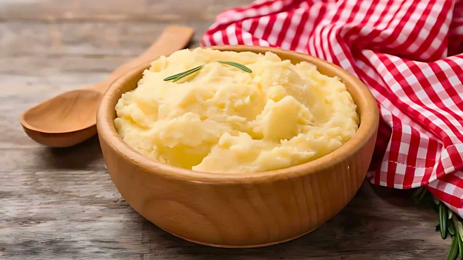 Bowl of mashed potatoes with a sprig of rosemary, next to a wooden spoon and a red and white checkered cloth on a wooden surface.